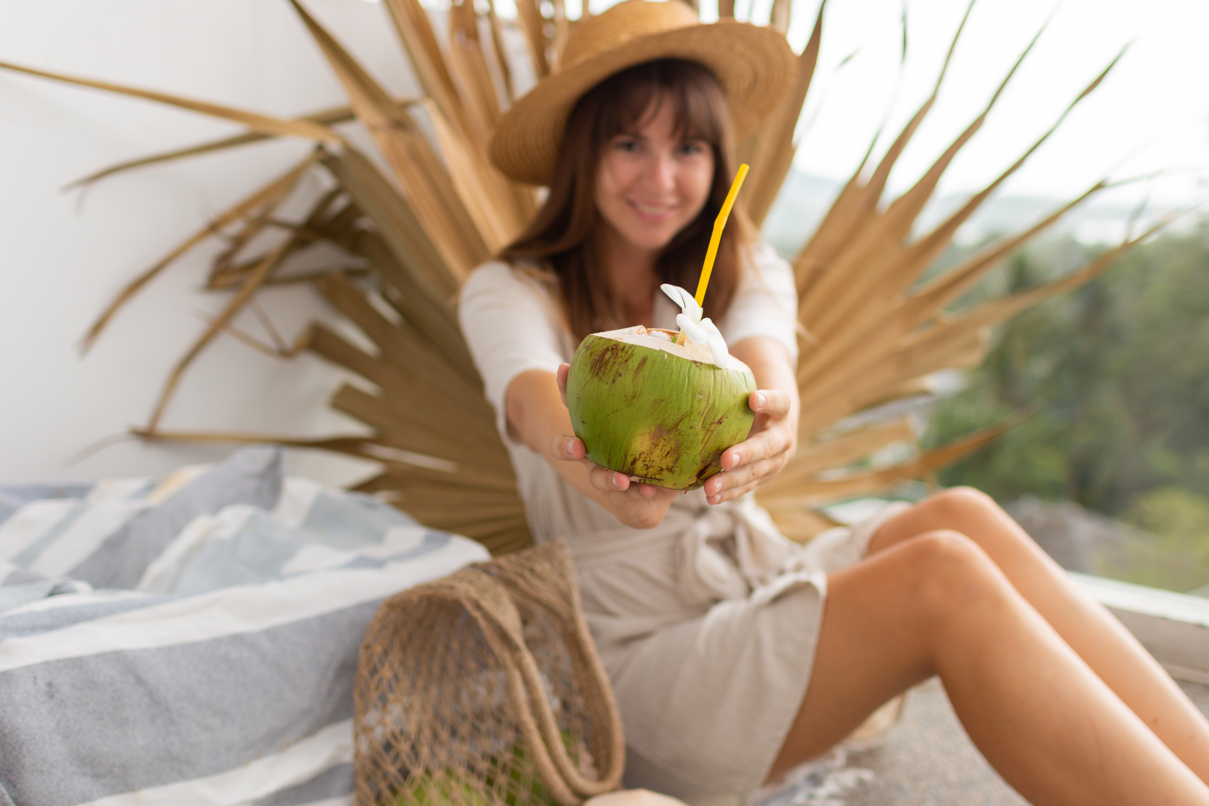 Pretty  brunette woman in straw hat  and linen dress posing  on terrace over dry palm  leaf holding Fresh coconut.